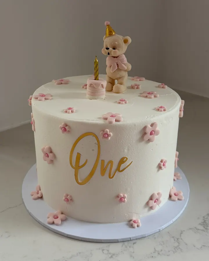 White first birthday cake dotted with little pink sugar flowers, a gold One and a teddy bear topper