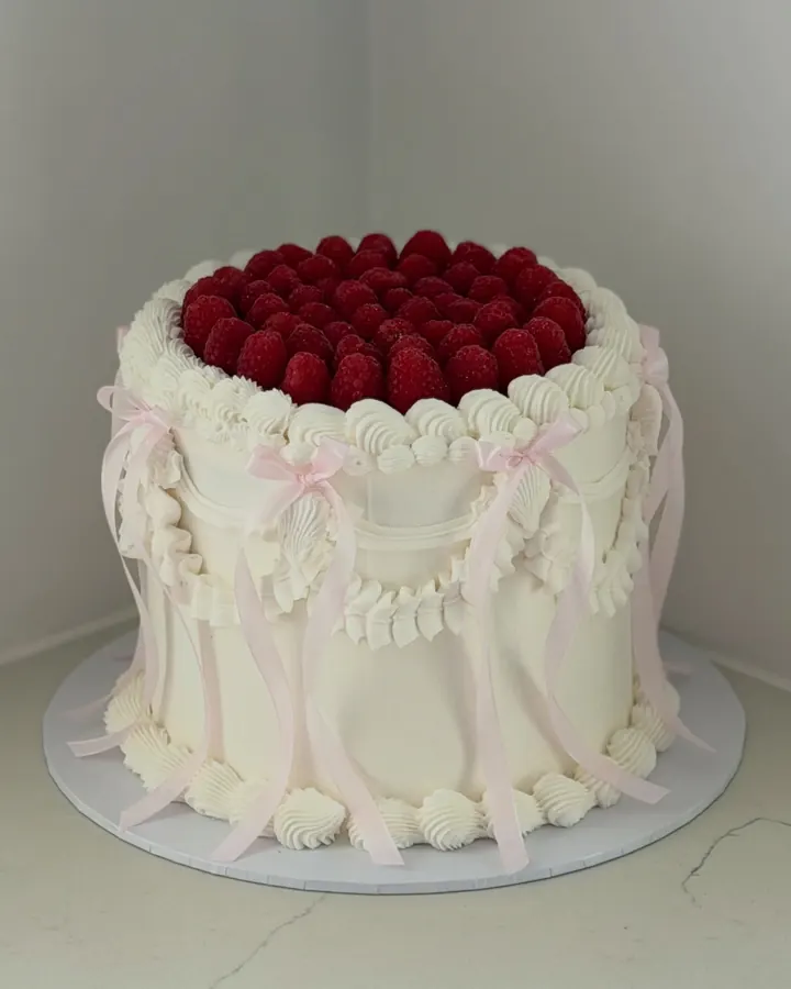 Tall white vintage cake with draped shell piping, pink ribbon bows and a crown of raspberries