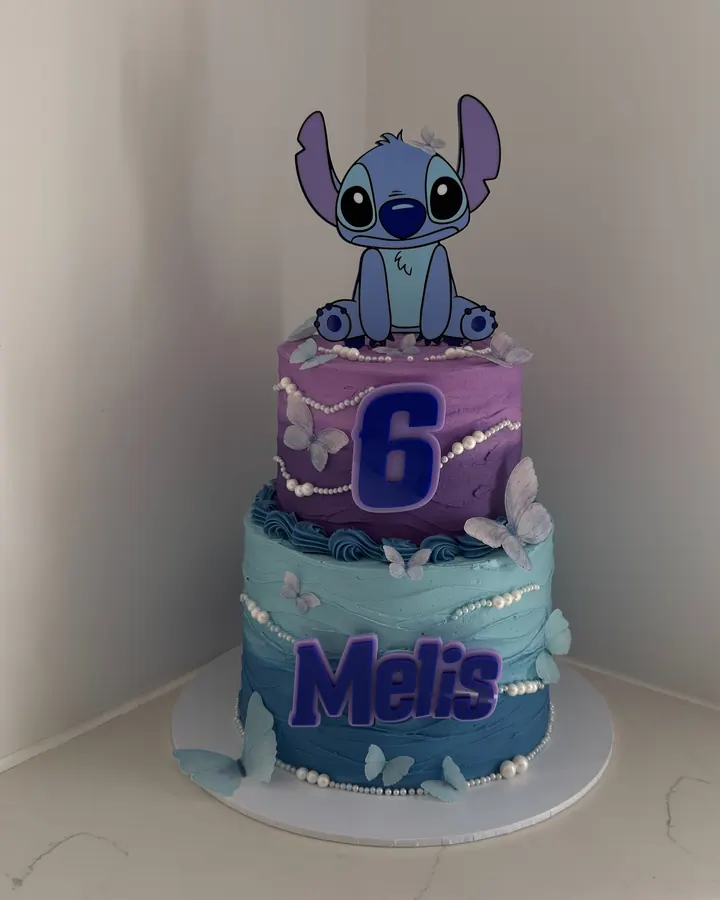 Two tier purple and blue Stitch cake with sugar butterflies, a number 6 and the name Melis